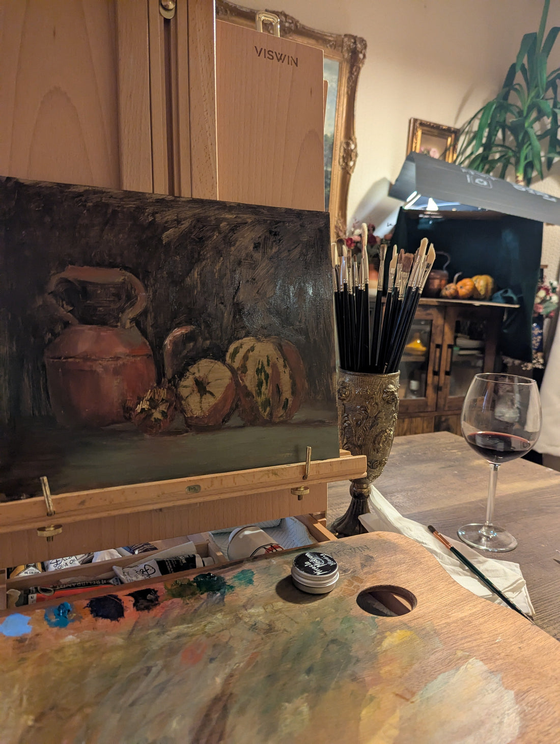 Oil painter's studio with still life painting on easel, palette in foreground and wine glass, brushes and the arranged diorama in the background. The main focal focus area for the painting artist.