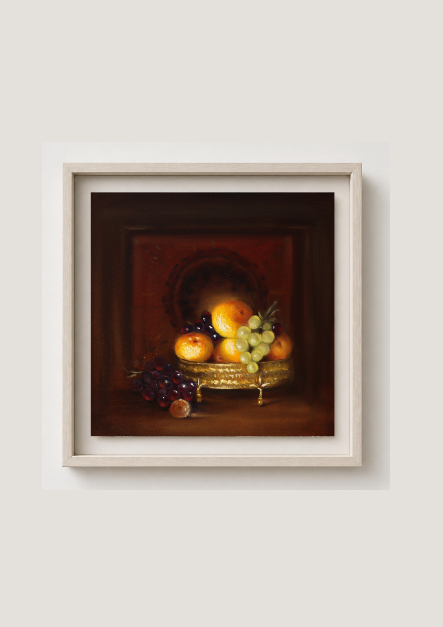 A framed still life painting of fruit, featuring oranges, grapes, and a pear on a gold tray, set against a dark brown background. The artwork is housed in a light beige frame and is mounted on a neutral light gray wall. Abundance - original chiaroscuro oil painting of a gilded fruit bowl with oranges and grapes by Marta Stolarska