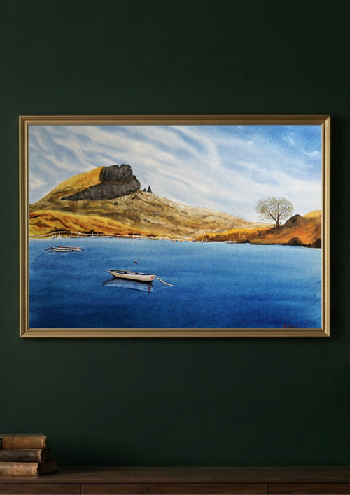 A framed landscape painting of a blue lake with boats, a grassy hill, and a tree under a cloudy sky, displayed on a dark green wall above a wooden surface.