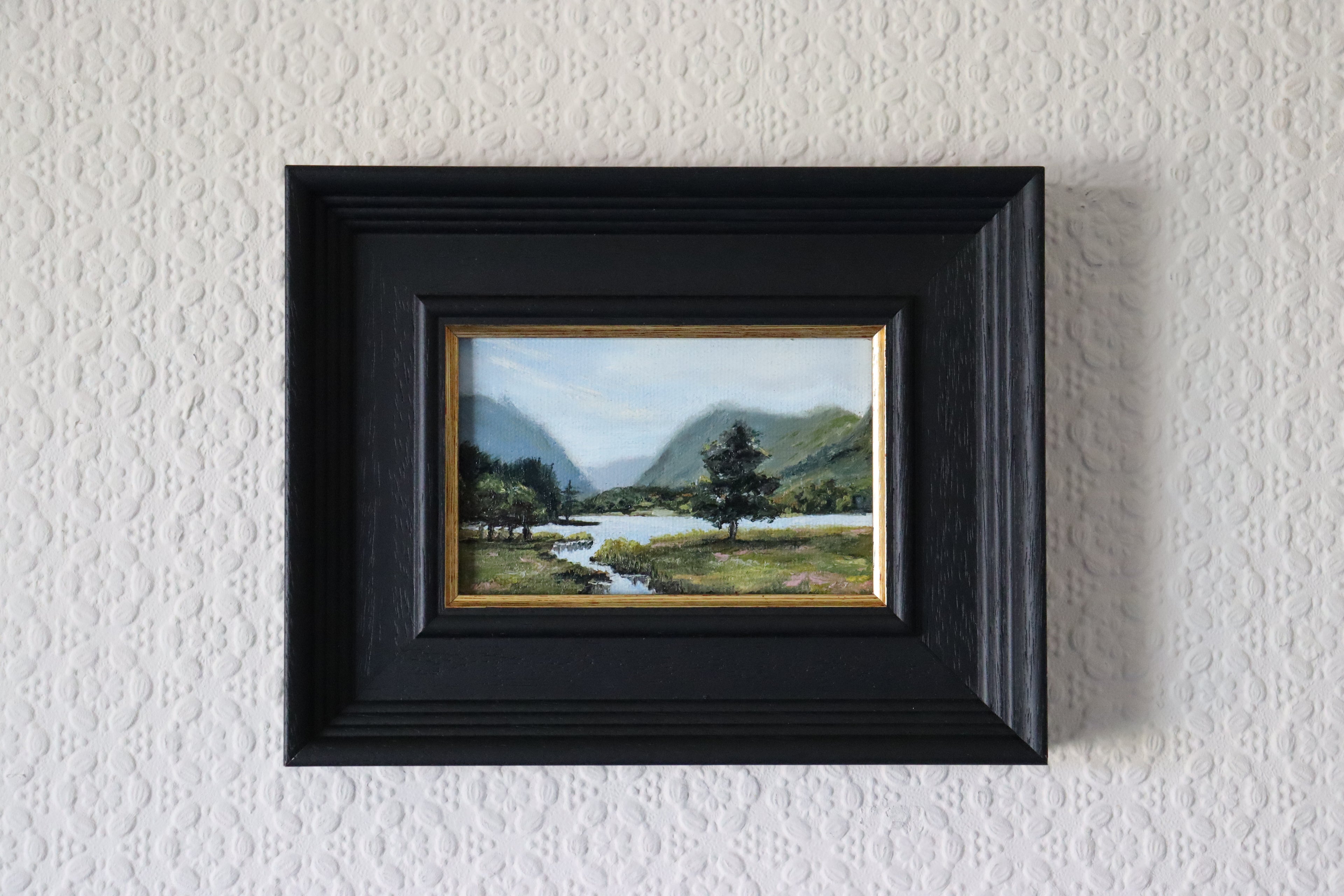 Framed painting of a landscape with a tree and river on a textured wall. Original oil Painting of a landscape with a stream, trees, and mountains.