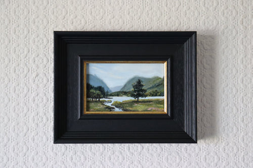 Framed painting of a landscape with a tree and river on a textured wall. Original oil Painting of a landscape with a stream, trees, and mountains.