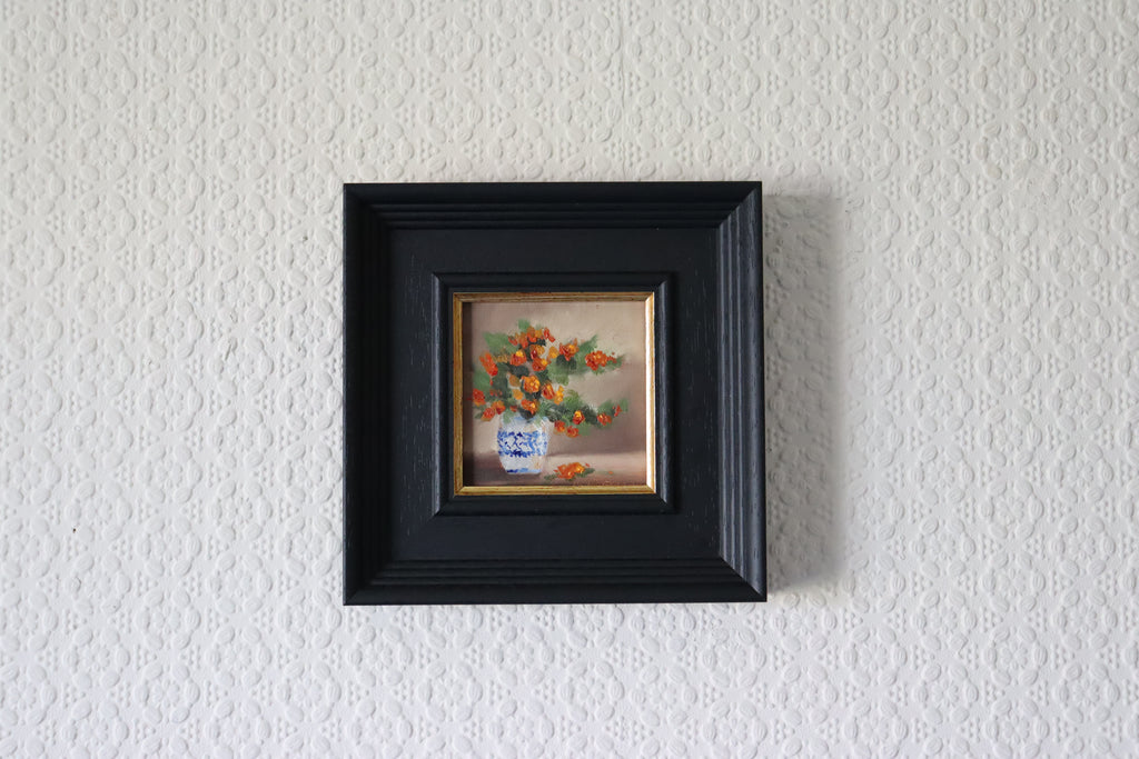 Small painting of flowers in a black frame on a textured white wall 