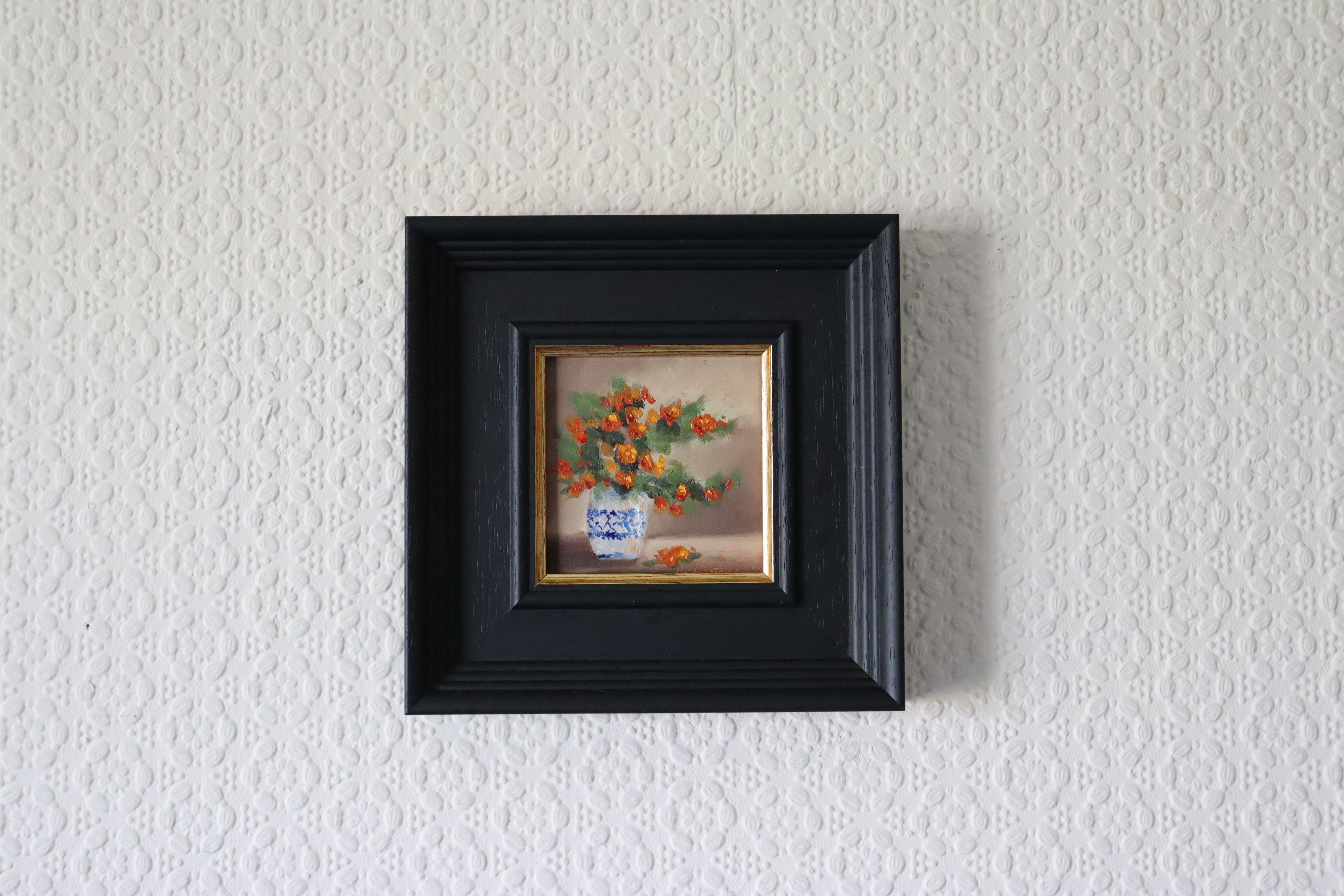 Small painting of flowers in a black frame on a textured white wall 