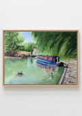 A framed watercolor painting of a canal scene with a blue and red narrowboat on green water, surrounded by trees and a stone quay, shown hanging on a light gray wall.