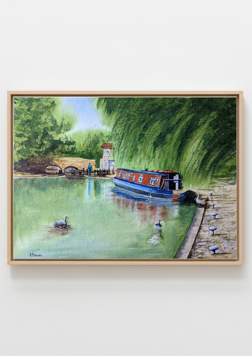 A framed watercolor painting of a canal scene with a blue and red narrowboat on green water, surrounded by trees and a stone quay, shown hanging on a light gray wall.