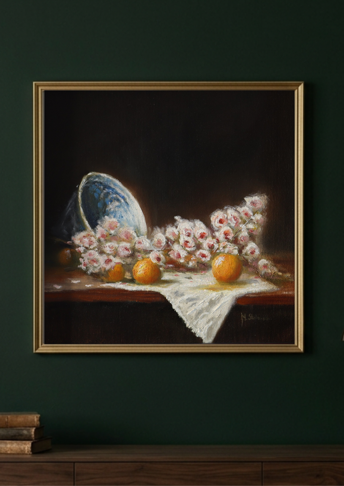 A framed still life painting hanging on a dark green wall above a wooden surface. The painting depicts pink and white flowers, two oranges, and a blue-and-white bowl on a dark background, enclosed in a gold frame.