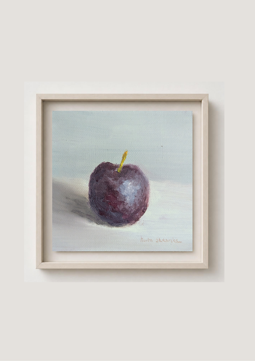 A framed still life painting of a dark purple plum with a small yellow stem, set against a pale blue-gray background. The artwork is housed in a light beige wooden frame and is mounted on a neutral light gray wall.