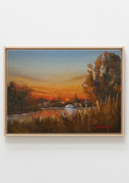 A framed landscape oil painting depicting a sunset over a river with trees and reeds, mounted on a light gray wall. Original oil painting of boaters enjoying sunset at the Suffolk marshes, warm evening light on water by local artist