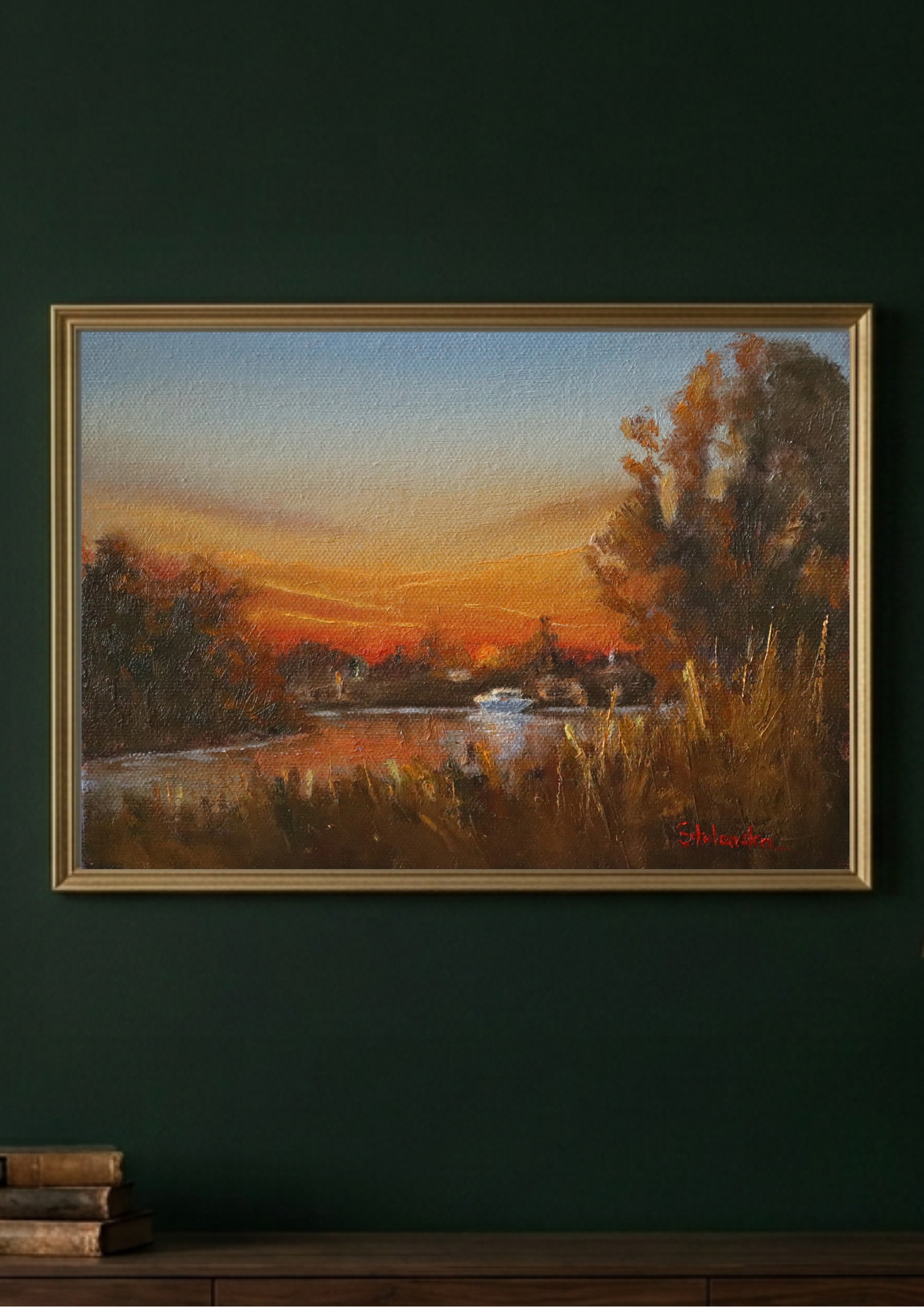 A framed landscape painting of a sunset over a river with trees and reeds, displayed on a dark green wall above a wooden surface with stacked books.