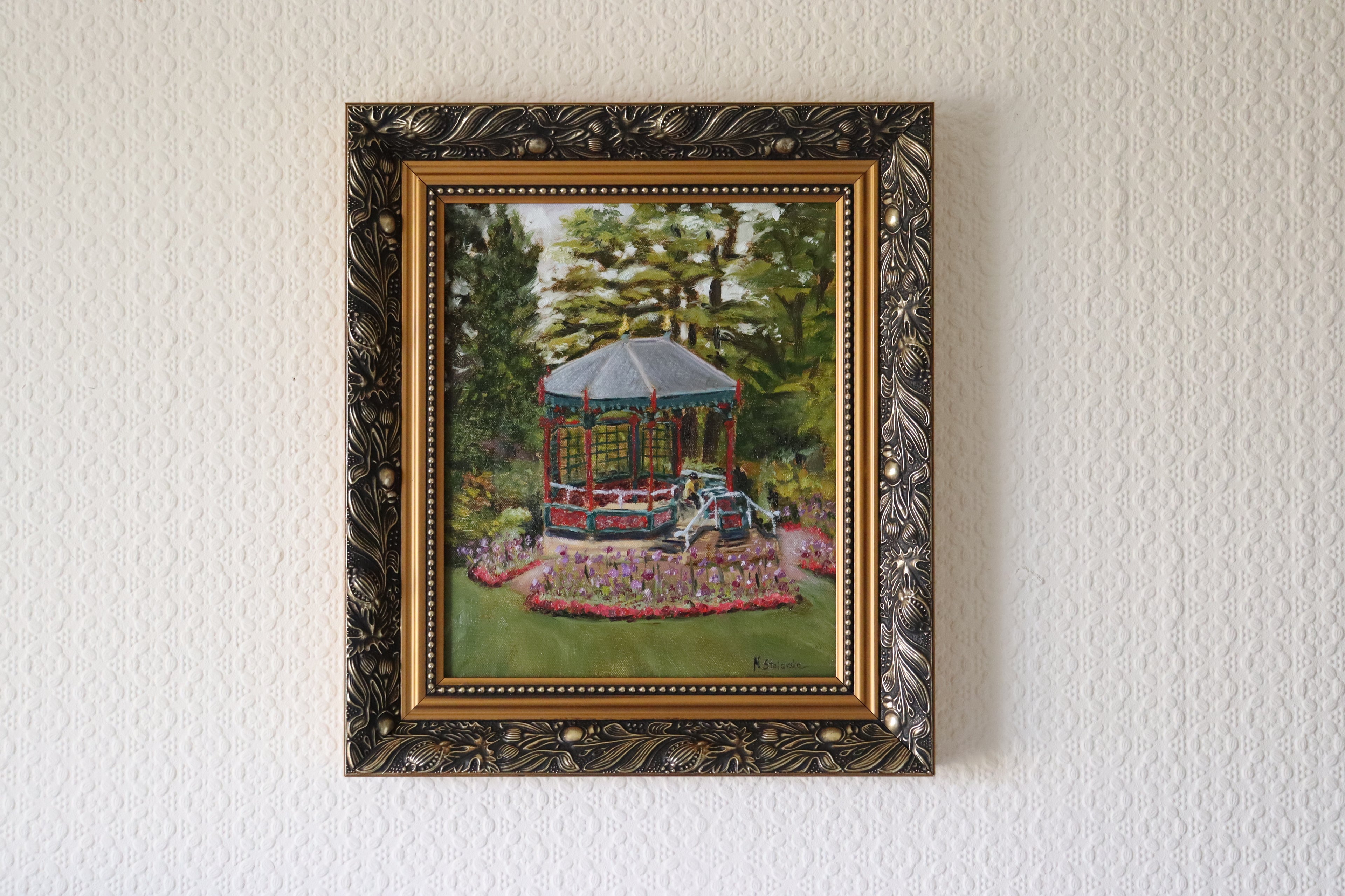 Framed in an ornate gold frame, Original oil painting done on location ( alla prima en plain air) at Birmingham Botanical Gardens. Depicts the band stand and a drawing artist with flowers and tress around Framed painting of a gazebo in a garden setting on a textured wall.