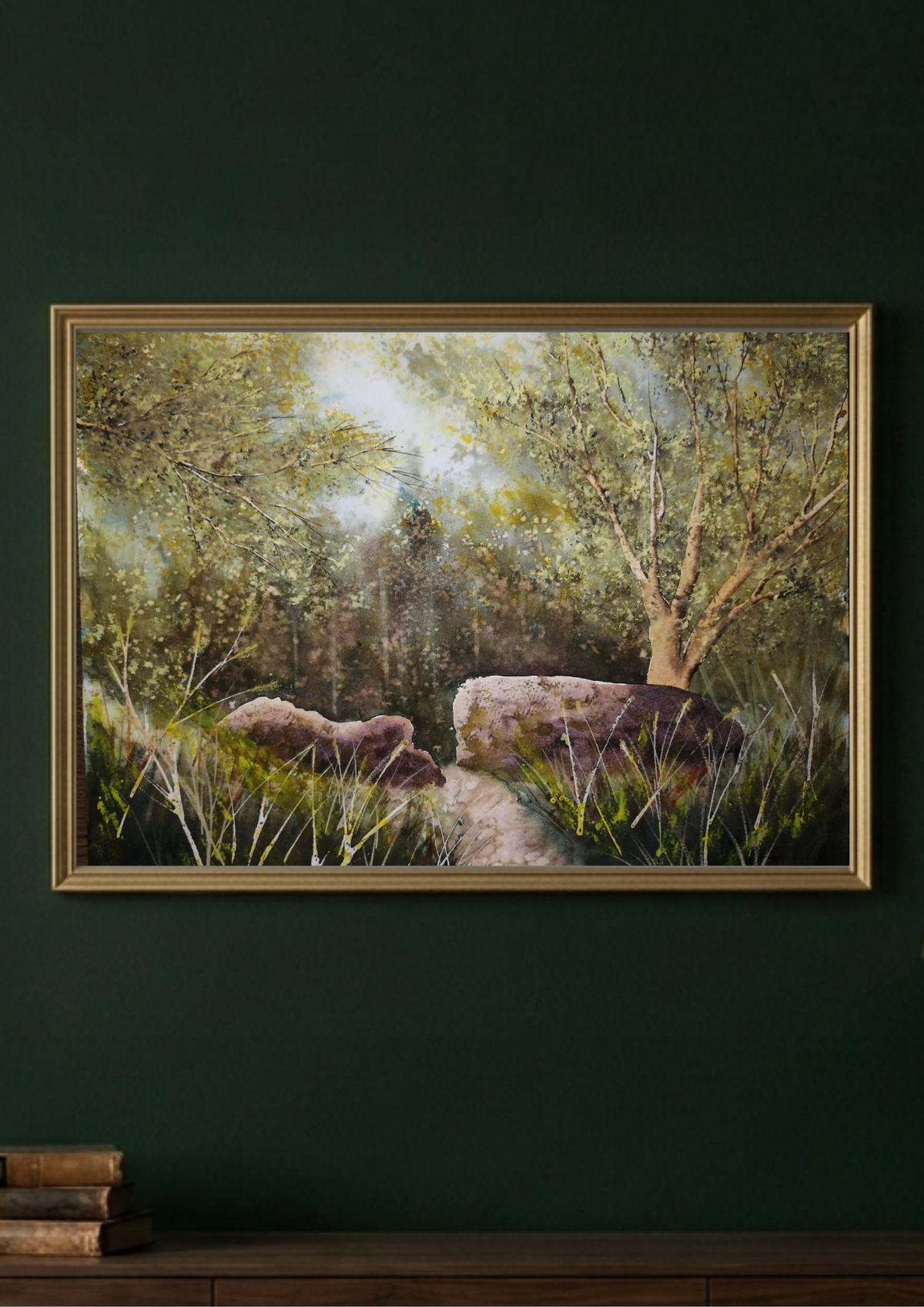 A framed landscape painting of a sunlit forest path with trees, rocks, and grass, displayed on a dark green wall above a wooden surface with stacked books.