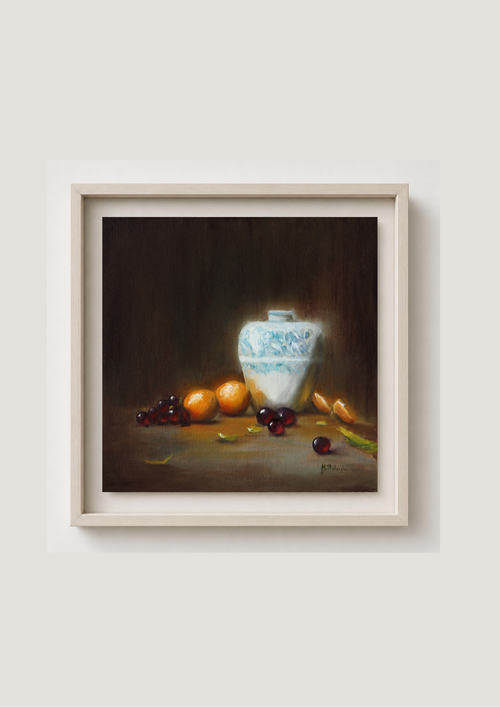 A framed still life painting featuring a white and blue patterned ceramic vase on a dark brown tabletop, surrounded by oranges, cherries, and a lemon, set against a light beige wall. Bouncing Light - original oil still life by Marta Stolarska, large white vase glowing with reflected light, oranges and grapes, painted from life on cradled wooden canvas panel, 30x30cm