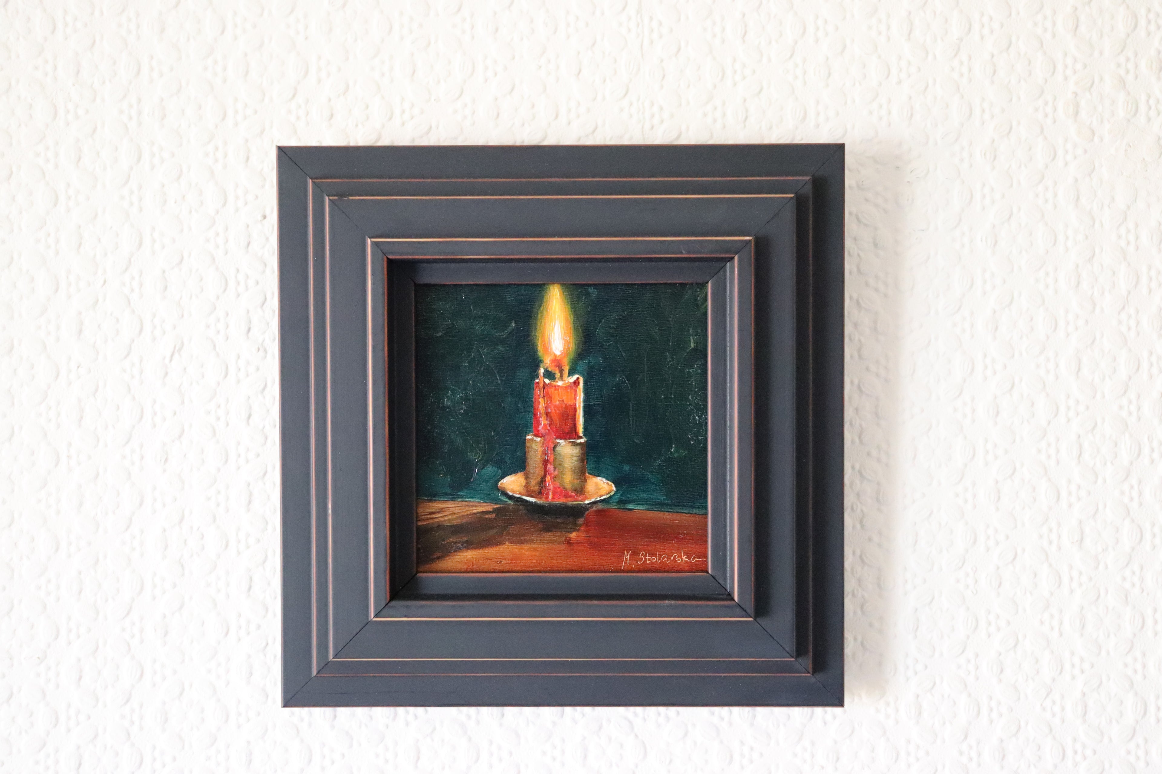 Framed painting of a candle on a textured wall. Original oil Painting of a lit red candle on a dark green background on a cradled panel. 
