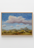 A framed landscape painting featuring a blue sky with large white and gray clouds, distant hills, and yellow-green trees in the foreground, shown against a plain white wall. 