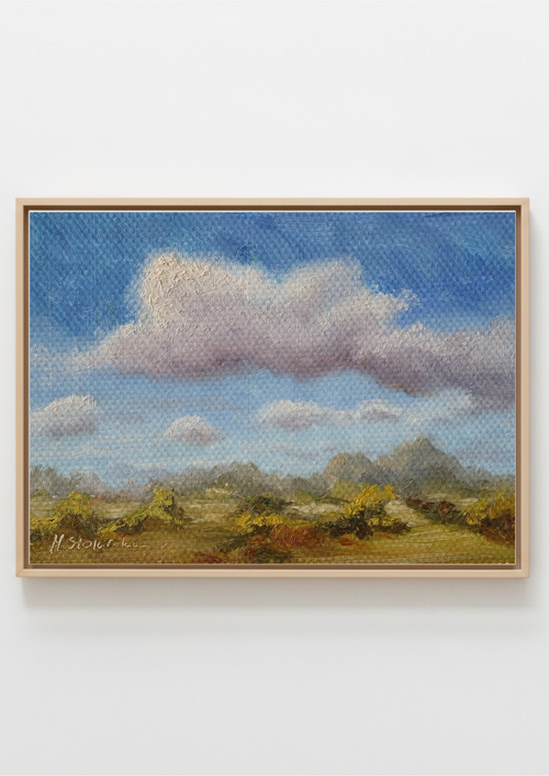 A framed landscape painting featuring a blue sky with large white and gray clouds, distant hills, and yellow-green trees in the foreground, shown against a plain white wall. 
