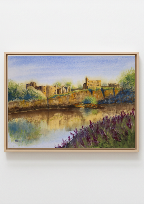 A framed watercolor painting of a scenic landscape featuring a river, stone ruins on a cliff, purple flowers in the foreground, and a blue sky, presented in a light wood frame against a white wall. Painting made by a local artist. Original watercolour painting of Chepstow Castle and cliffs from across the River Wye, Wales, Britain's oldest stone fortress. Watercolor painting of a castle by a lake with purple flowers in the foreground.