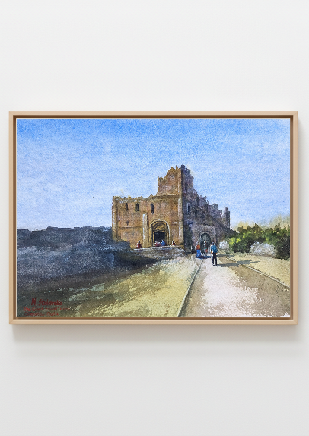 A framed watercolor painting of a historic stone archway or gatehouse under a blue sky, with a path in the foreground and small figures near the entrance, shown against a light neutral wall. Painting of a castle with people walking towards it on a path. Original watercolour painting of Chepstow Castle great tower viewed from within the castle grounds, Wales