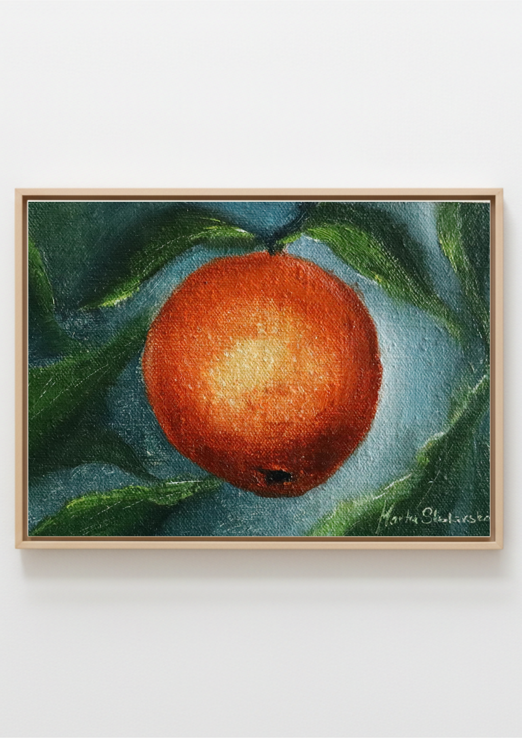 A framed still life painting of a vibrant orange with green leaves, presented in a light wood frame against a clean white wall. Childhood Scents - original oil still life by Marta Stolarska, a single orange with leaves on a dark background, painted on handmade linen board, 10 x 15 cm