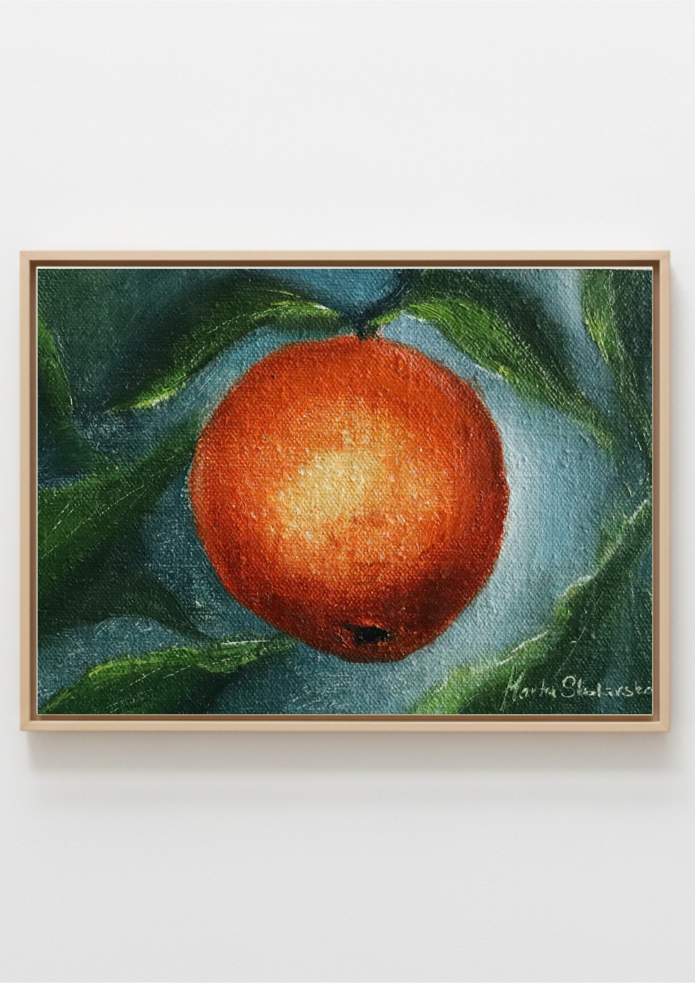 A framed still life painting of a vibrant orange with green leaves, presented in a light wood frame against a clean white wall. Childhood Scents - original oil still life by Marta Stolarska, a single orange with leaves on a dark background, painted on handmade linen board, 10 x 15 cm