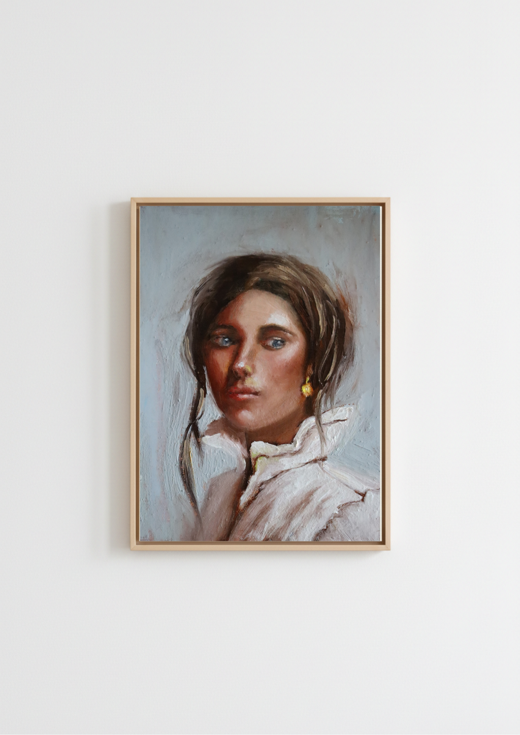 A framed portrait painting of a young woman with brown hair, blue eyes, and a white ruffled collar, hanging on a plain white wall. Composed - original oil portrait by Marta Stolarska, dark-haired woman with blue eyes and glowing skin in white high-collar shirt with gold dangling earring, oil on canvas board, Subtle Expressions collection