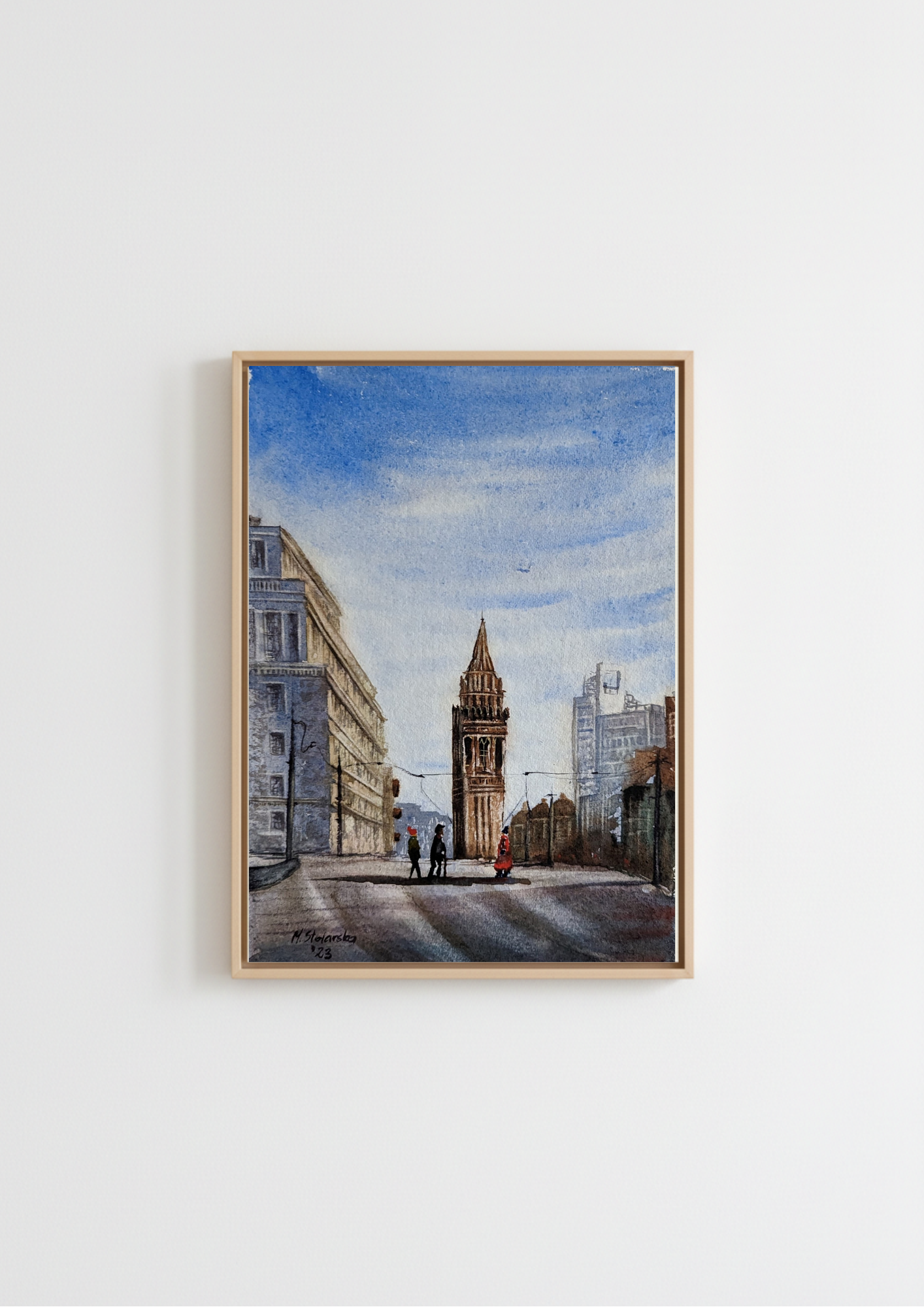 A framed watercolor painting of a city street scene with a prominent tower, hanging on a plain white wall. Corporation Street in Winter Birmingham - limited edition fine art print by Marta Stolarska, tram lines and silhouetted figures crossing the street, based on original watercolour, A4 archival matt paper, edition of 50, hand signed and numbered