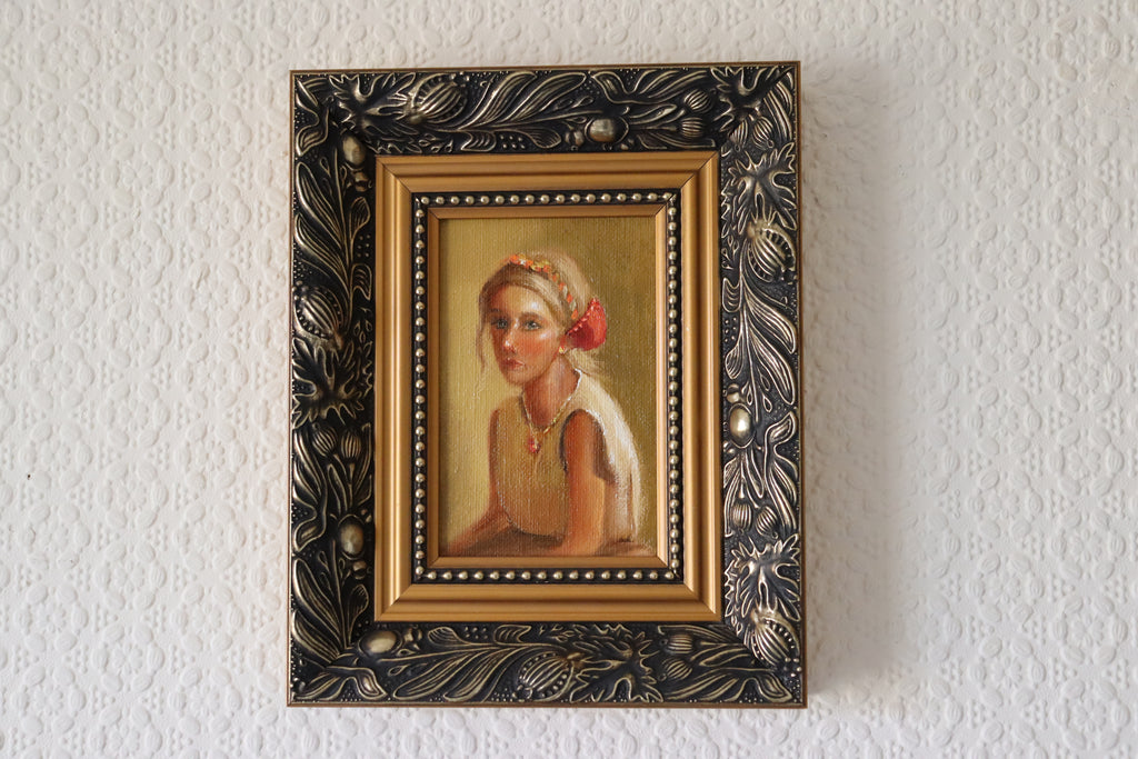 Framed lifestyle setting of an Original oil portrait painting of a golden-haired girl in a linen dress with headband, direct emotive gaze, framed in gold