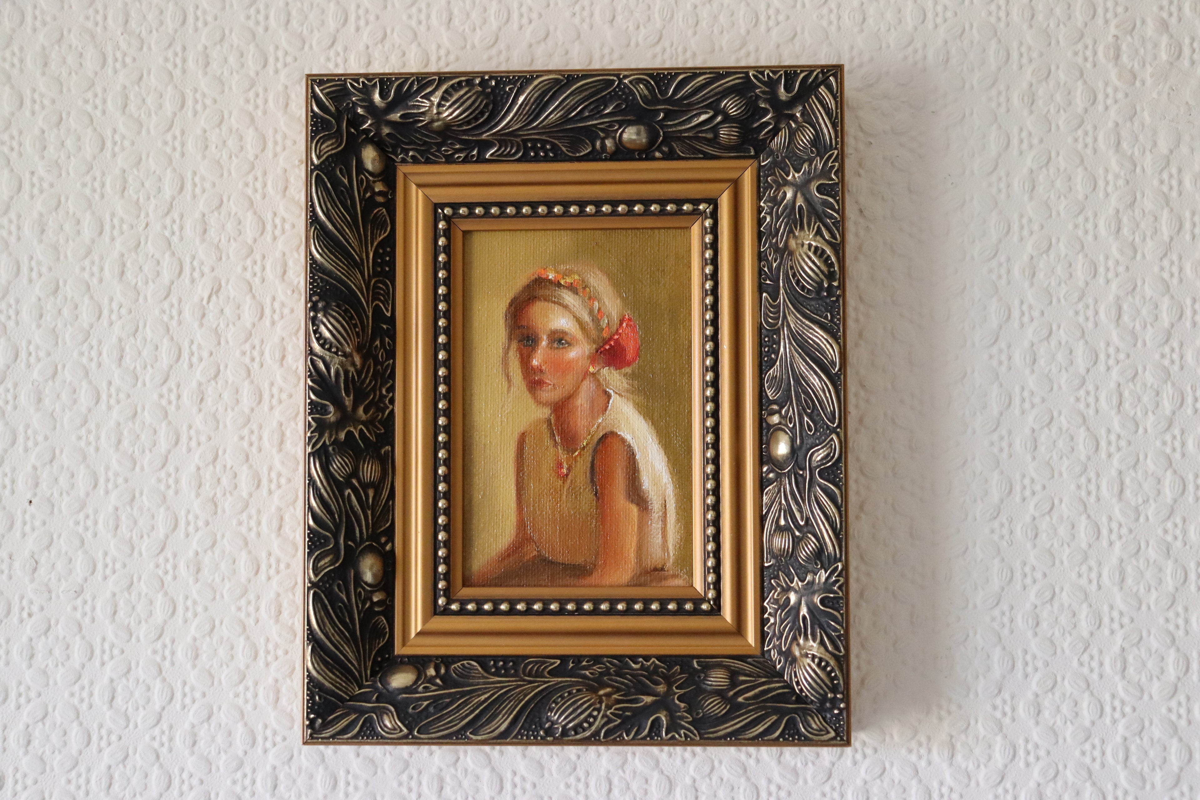Framed lifestyle setting of an Original oil portrait painting of a golden-haired girl in a linen dress with headband, direct emotive gaze, framed in gold