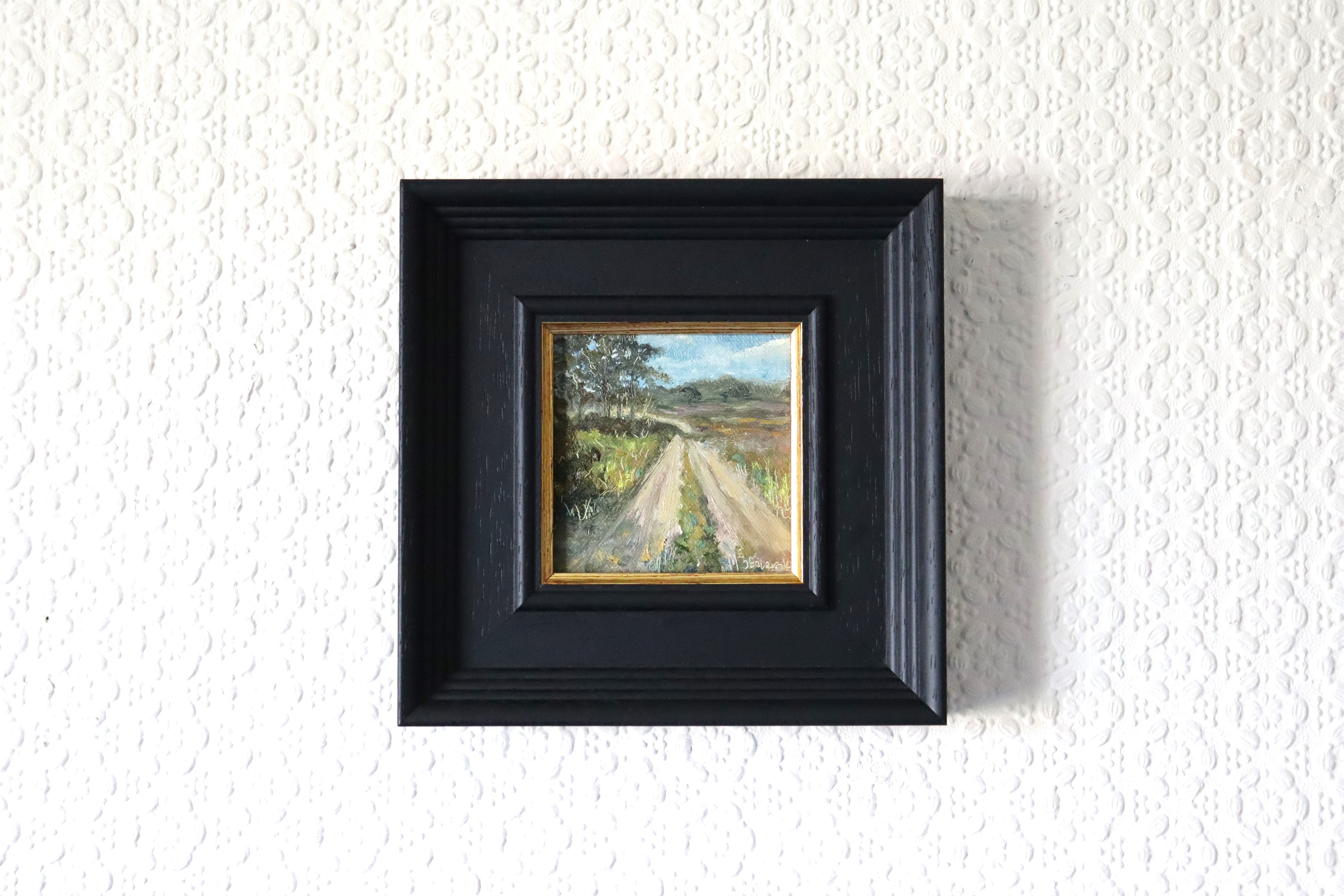 Framed painting of a landscape with a path on a textured white wall. Lifestyle photo of an Original oil painting of a country road leading into open fields, simple English countryside scene framed