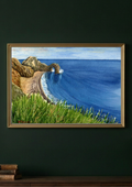 A framed landscape painting of a coastal scene with blue ocean water, rocky cliffs, and green grass, displayed on a dark green wall above a wooden surface.