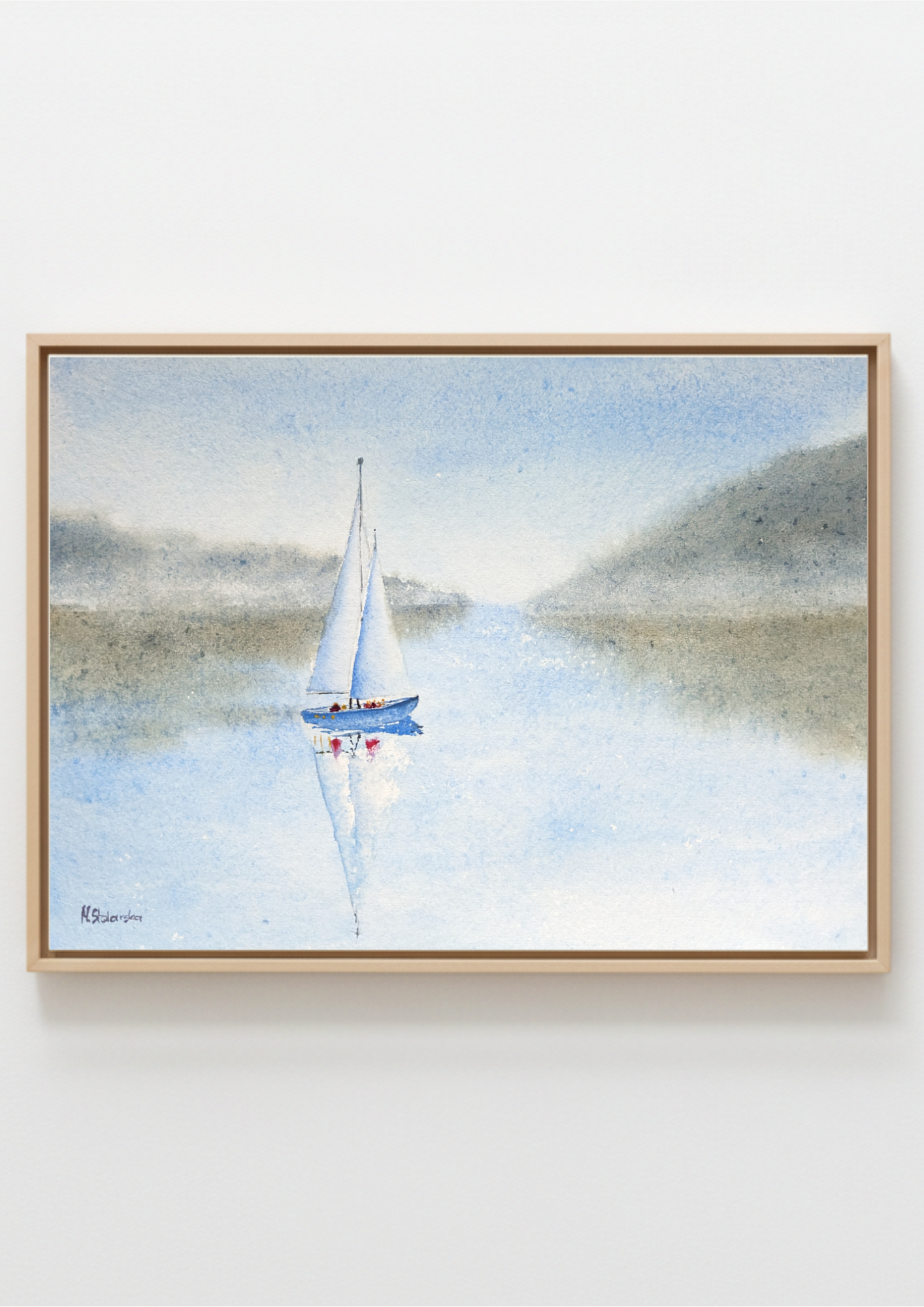 A framed watercolor painting of a sailboat on a calm lake with misty hills in the background, presented in a light wood frame against a white wall.
