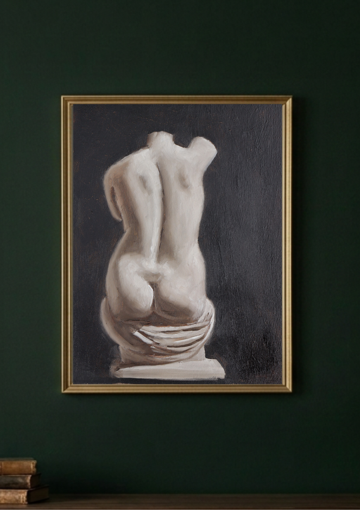 A framed original art featuring a classical sculpture of a draped female torso, shown hanging on a dark green wall above a wooden surface with books.