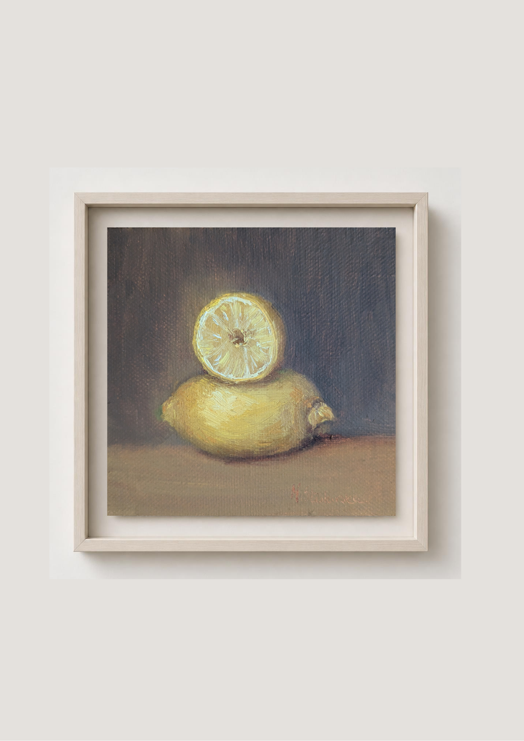 A framed still life painting of a yellow lemon with a sliced half on top, set against a dark gray background, enclosed in a light beige frame and mounted on a neutral light gray wall.
