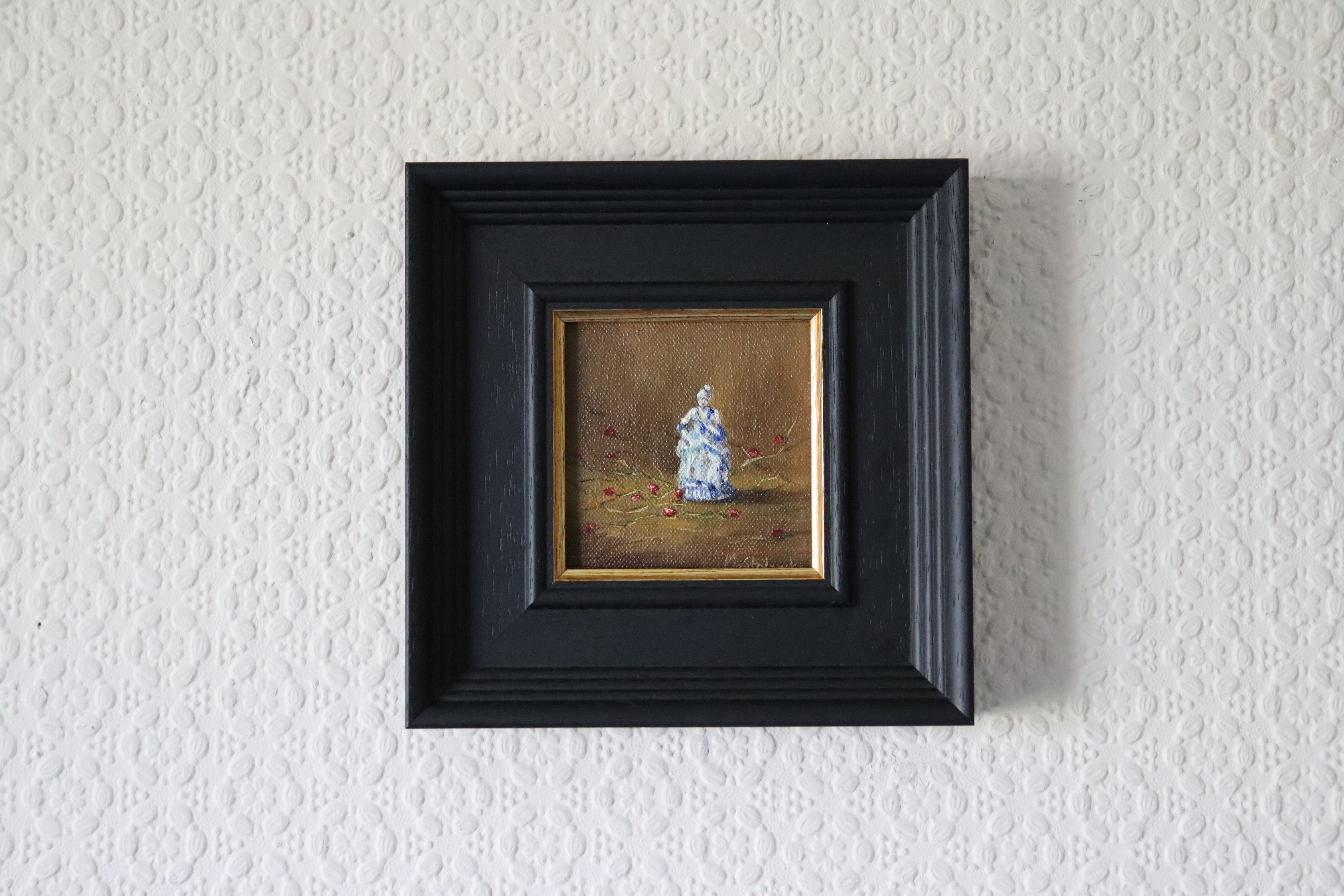 Small painting of a figure in a black frame on a textured white wall. Original oil painting Frozen in Time, still life capturing a moment of quiet beauty and stillness