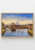 A framed landscape art print depicting a canal scene with boats, bridges, and buildings under a blue sky, mounted on a light gray wall.