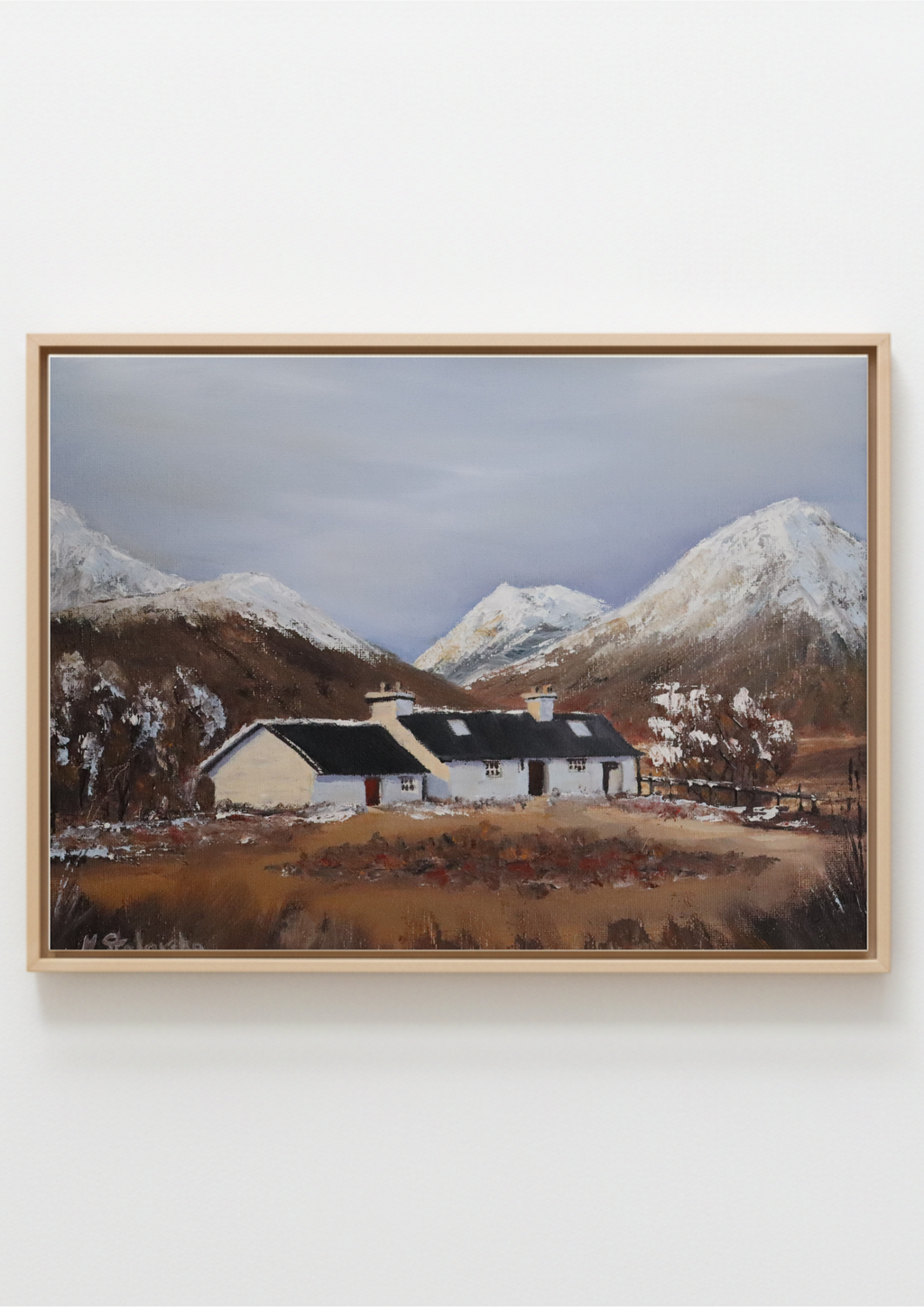 A framed landscape painting of a white cottage with a dark roof nestled in a valley, with snow-dusted mountains in the background under a cloudy sky, displayed on a light gray wall.