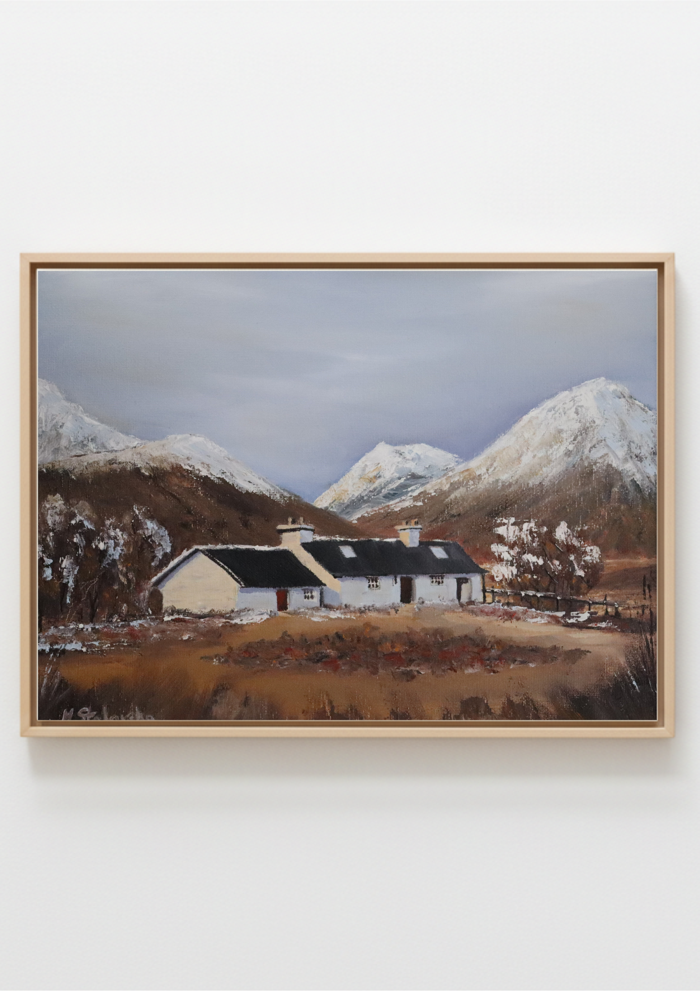 A framed landscape painting of a white cottage with a dark roof nestled in a valley, with snow-dusted mountains in the background under a cloudy sky, displayed on a light gray wall.