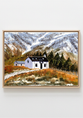 A framed landscape painting of a white cottage with a dark roof situated in a grassy field, with snow-dusted mountains in the background, shown hanging on a light gray wall. Original watercolour painting of Lagangarbh Cottage Glencoe in autumn, snow-capped Buachaille Etive Mor behind, framed