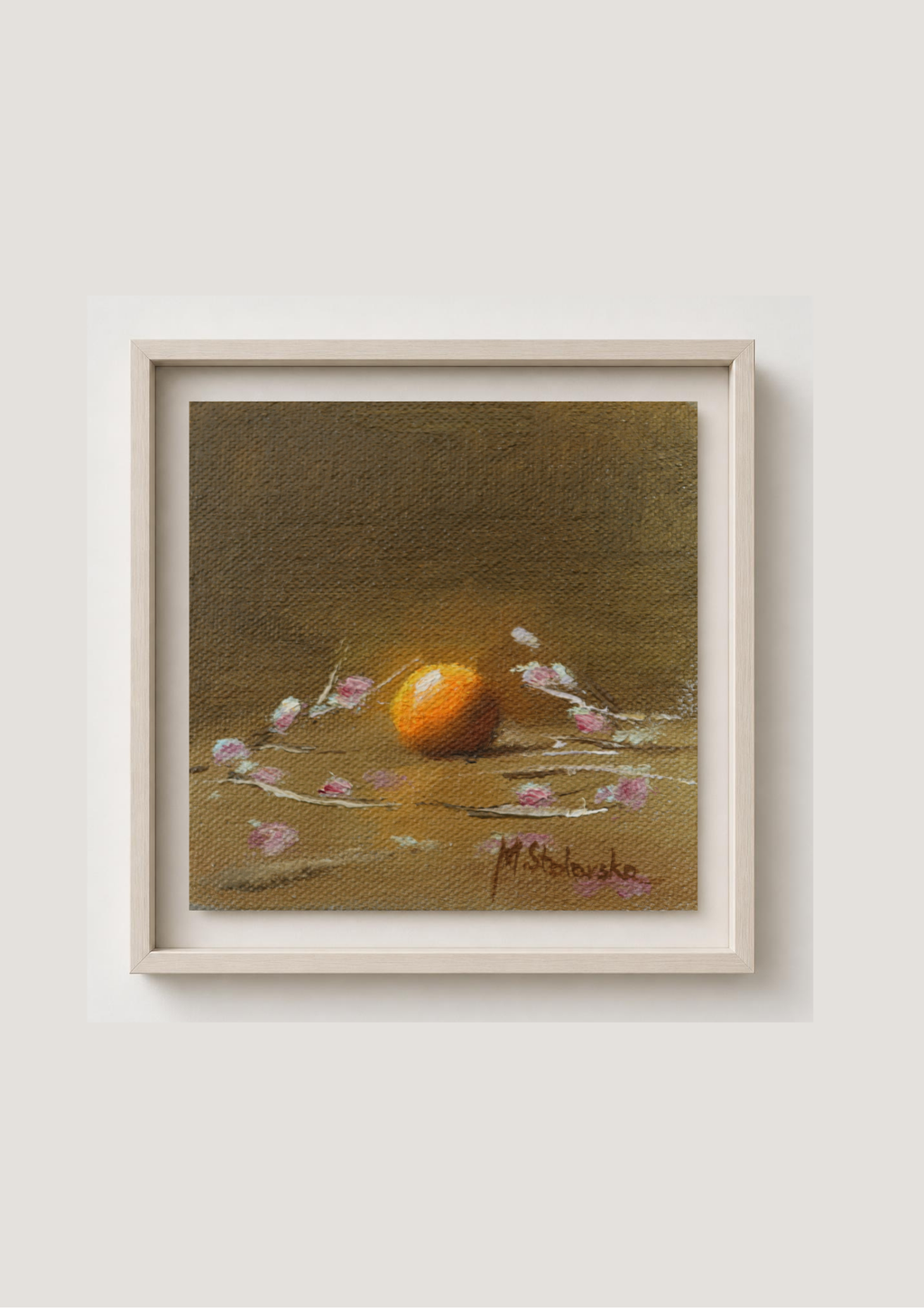 A framed still life painting featuring an orange surrounded by delicate pink blossoms on a textured brown background, housed in a light-colored wooden frame against a neutral wall.