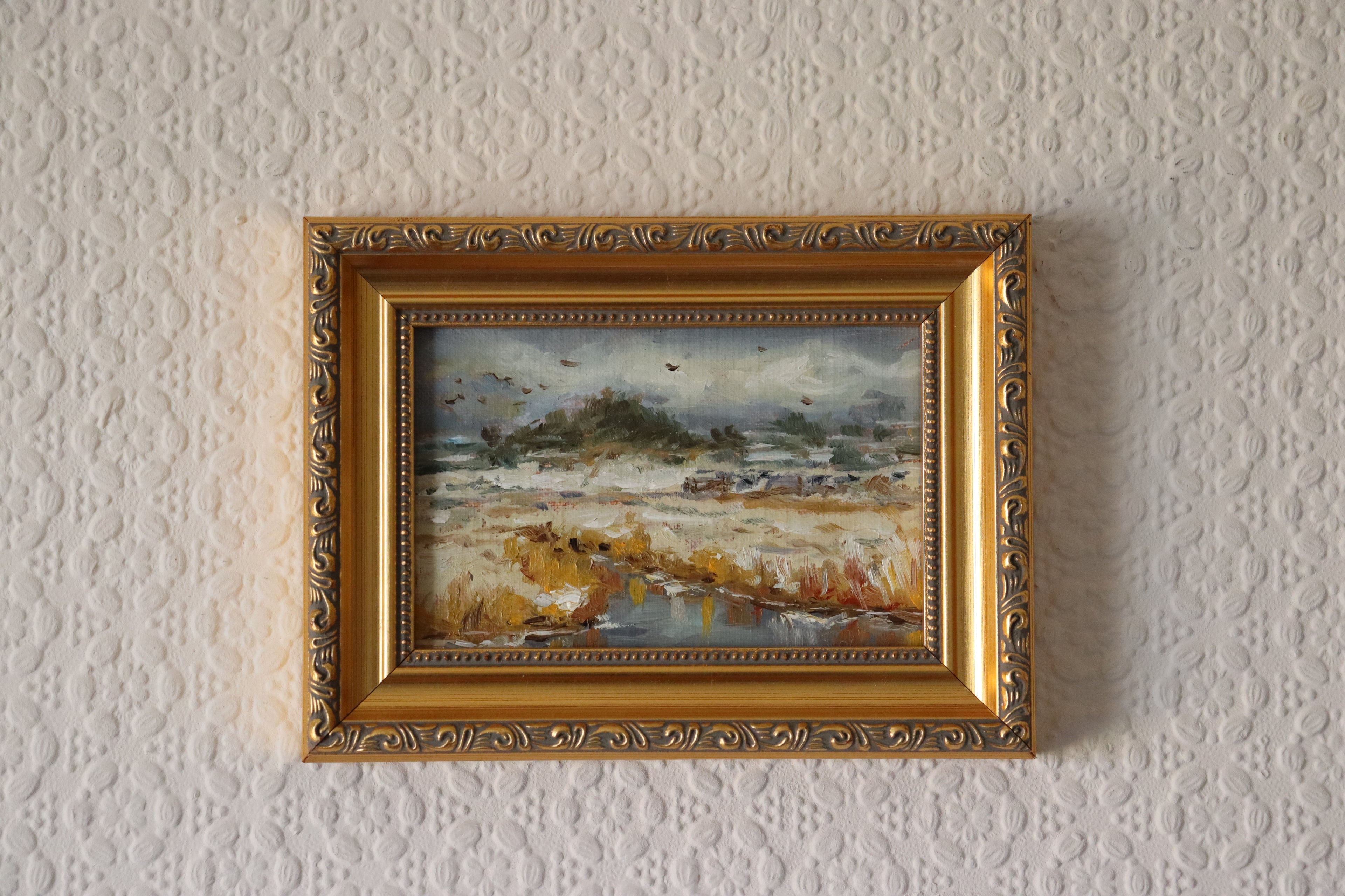 Framed painting of a winter landscape on a textured wall. Original oil painting of a moody Suffolk marsh, overcast atmospheric sky reflected in still water