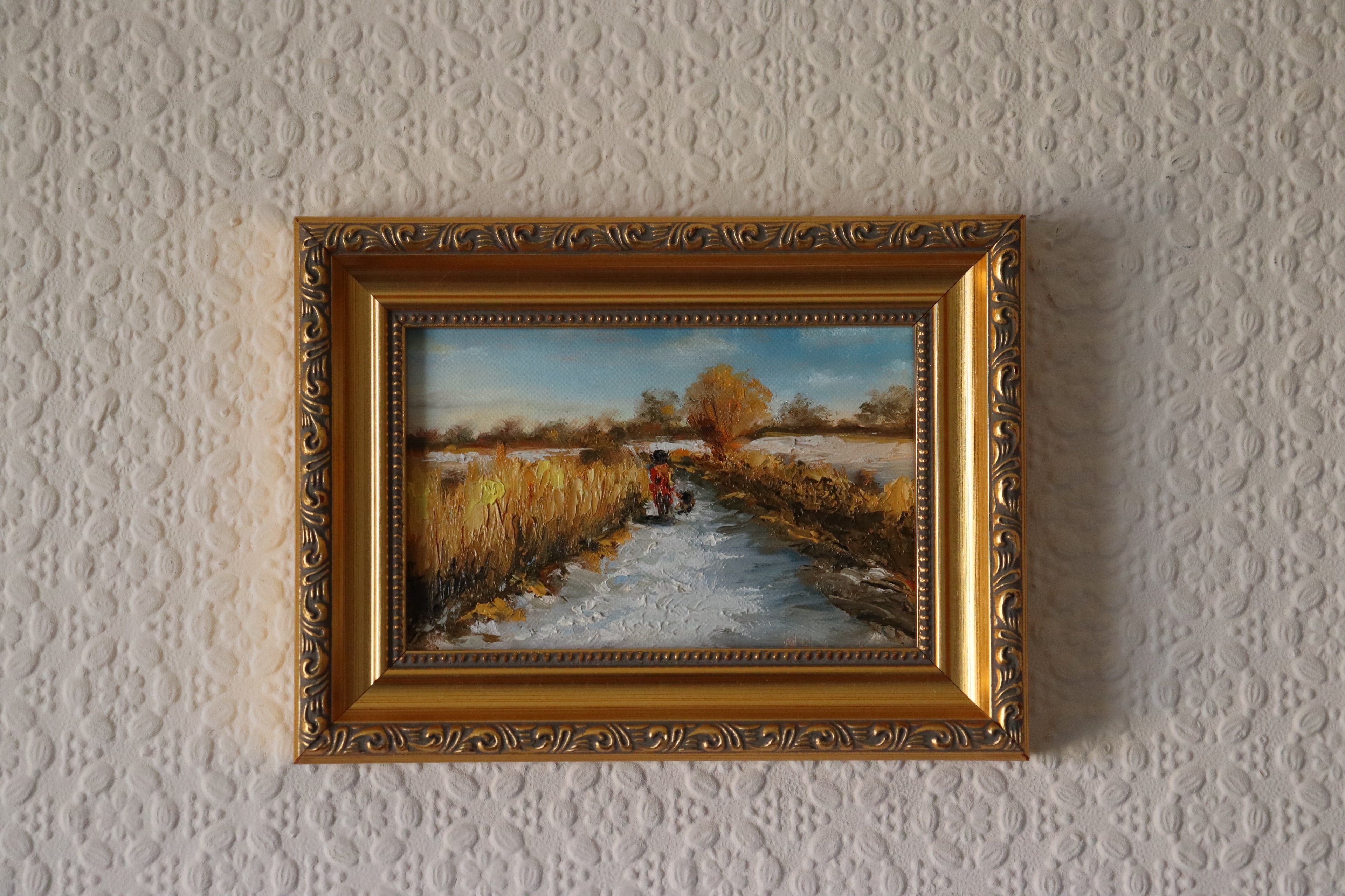 Painting of a rural scene with a person on a bike and a dog, surrounded by fields and trees by a local artist. Original oil painting of a winter walk at Suffolk marshes, snow-covered landscape with figures