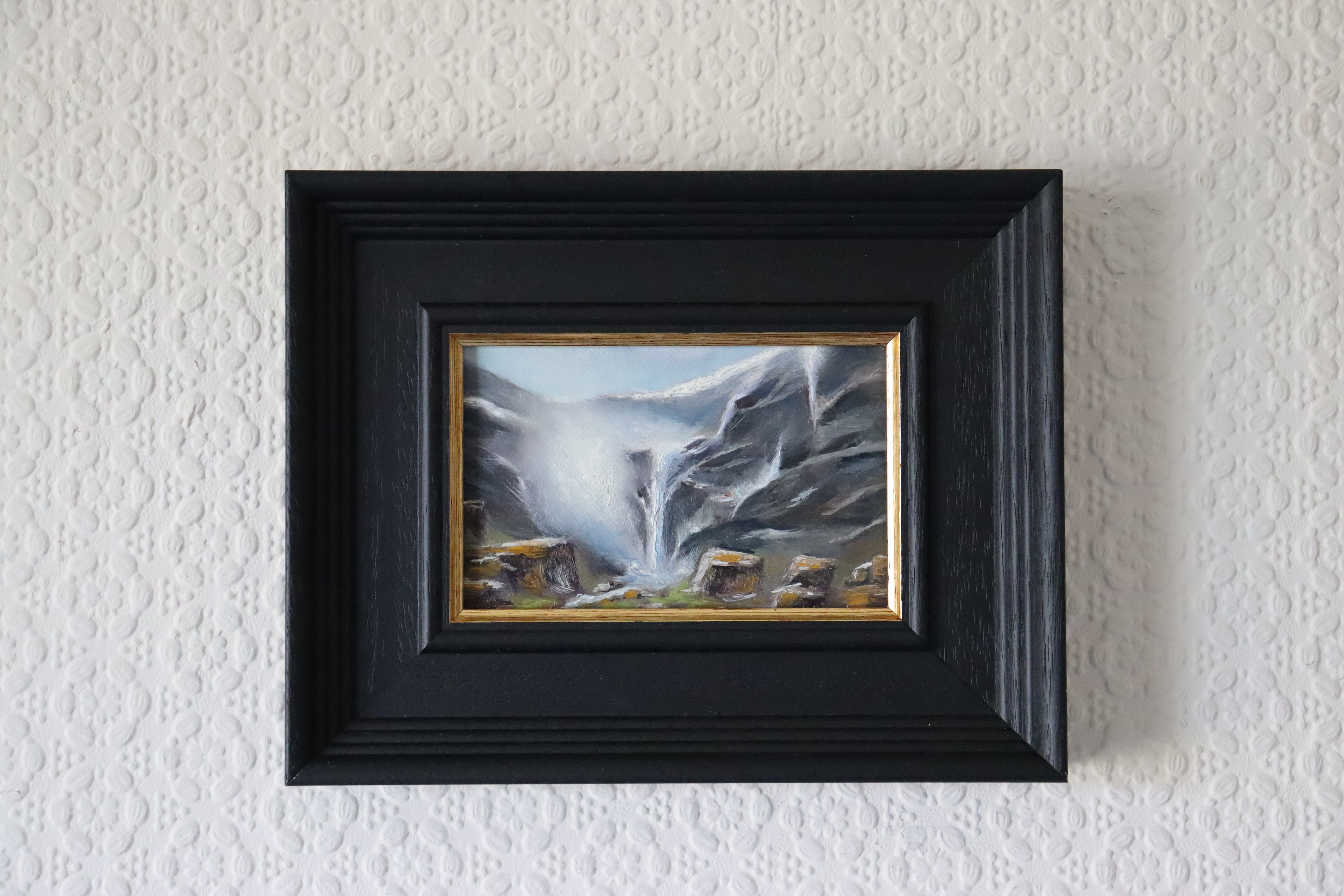 Framed, lifestyle image of an Original oil painting of an icy waterfall with mist and spray, dramatic winter landscape, black frame with gold bevel