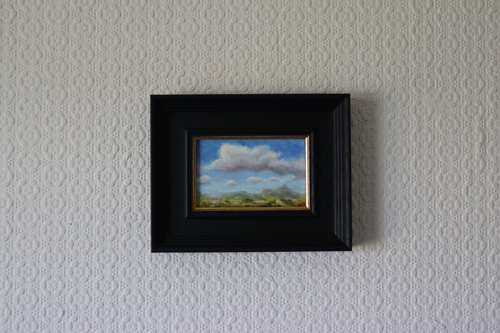 Framed painting of a landscape with clouds on a textured wall. Original oil painting of changing weather over open landscape, dramatic sky with clouds and shifting light