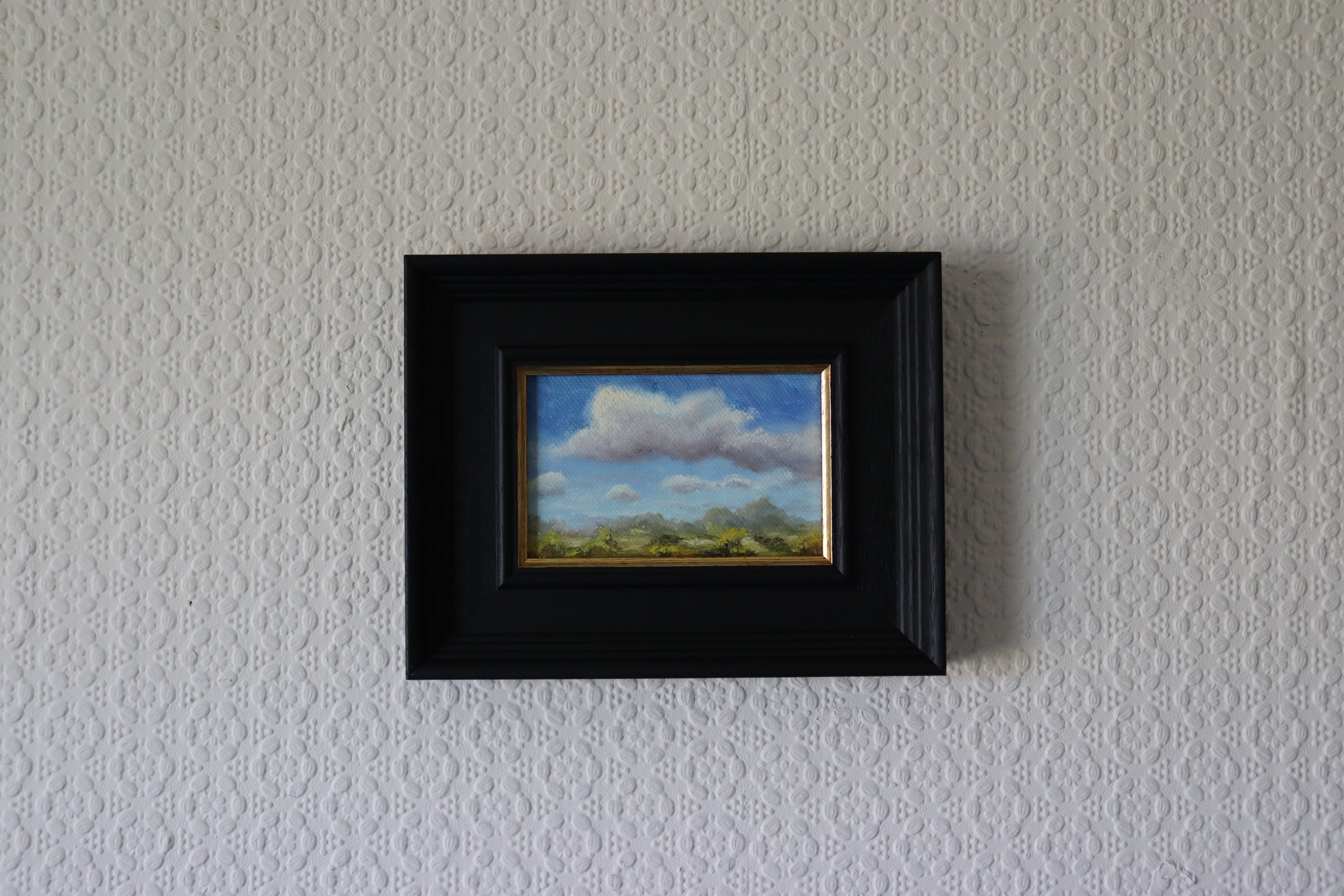 Framed painting of a landscape with clouds on a textured wall. Original oil painting of changing weather over open landscape, dramatic sky with clouds and shifting light