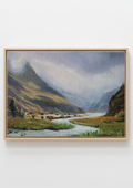 A framed landscape painting of a mountain valley with a winding river, shown against a plain white wall. The painting features green fields, dark rocky mountains, and a cloudy sky, enclosed in a light wood frame.