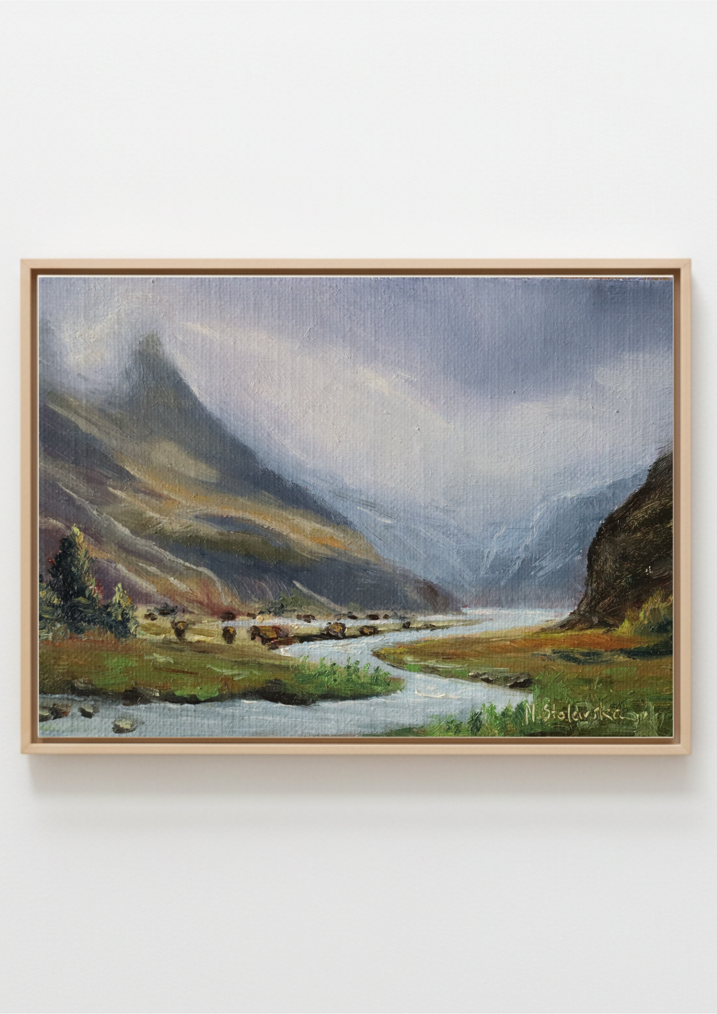 A framed landscape painting of a mountain valley with a winding river, shown against a plain white wall. The painting features green fields, dark rocky mountains, and a cloudy sky, enclosed in a light wood frame.