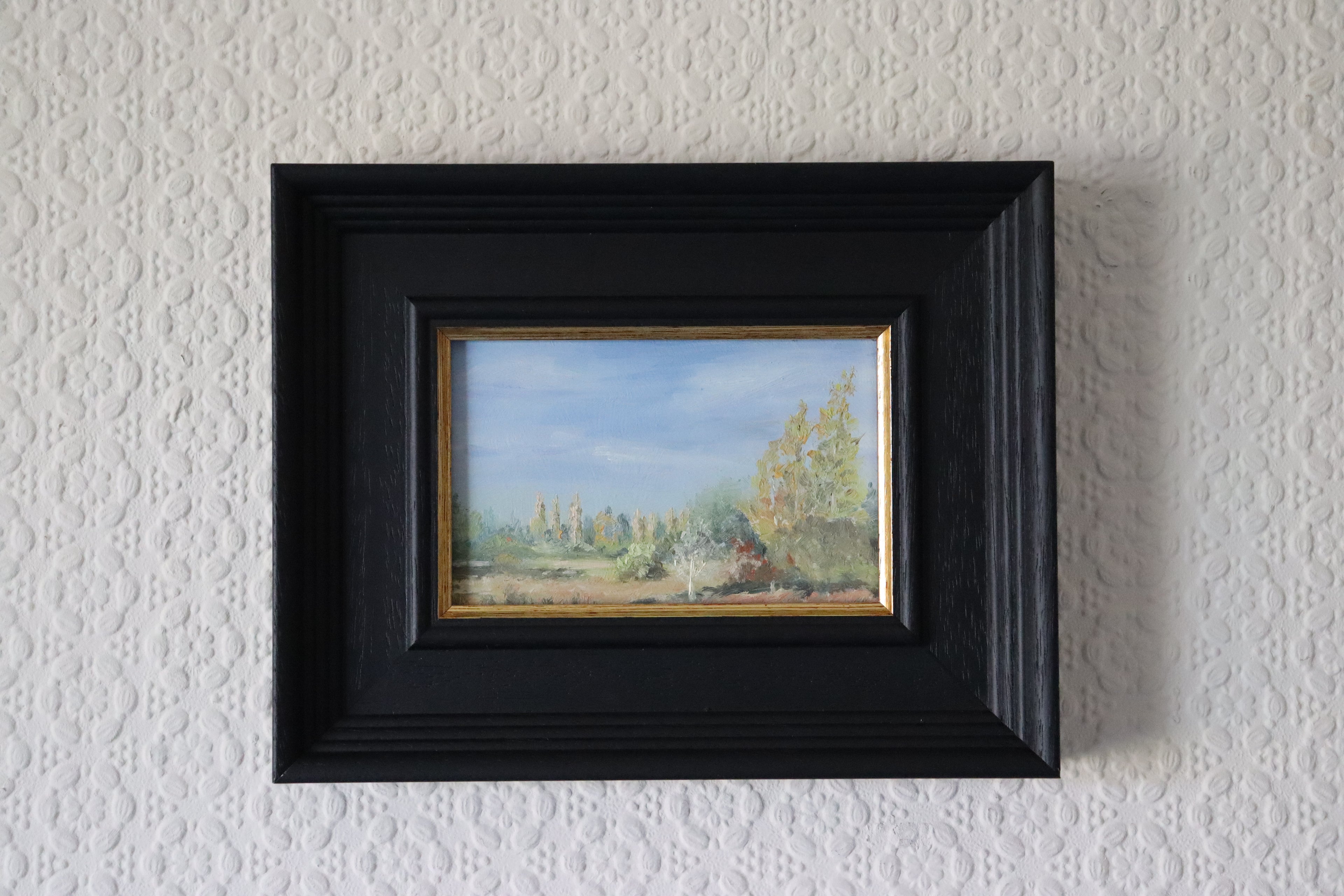 Framed painting of a landscape on a textured white wall. Scenic landscape with trees and a blue sky. Original oil painting of a path towards the meadows at Carlton Marshes Suffolk, and open fields