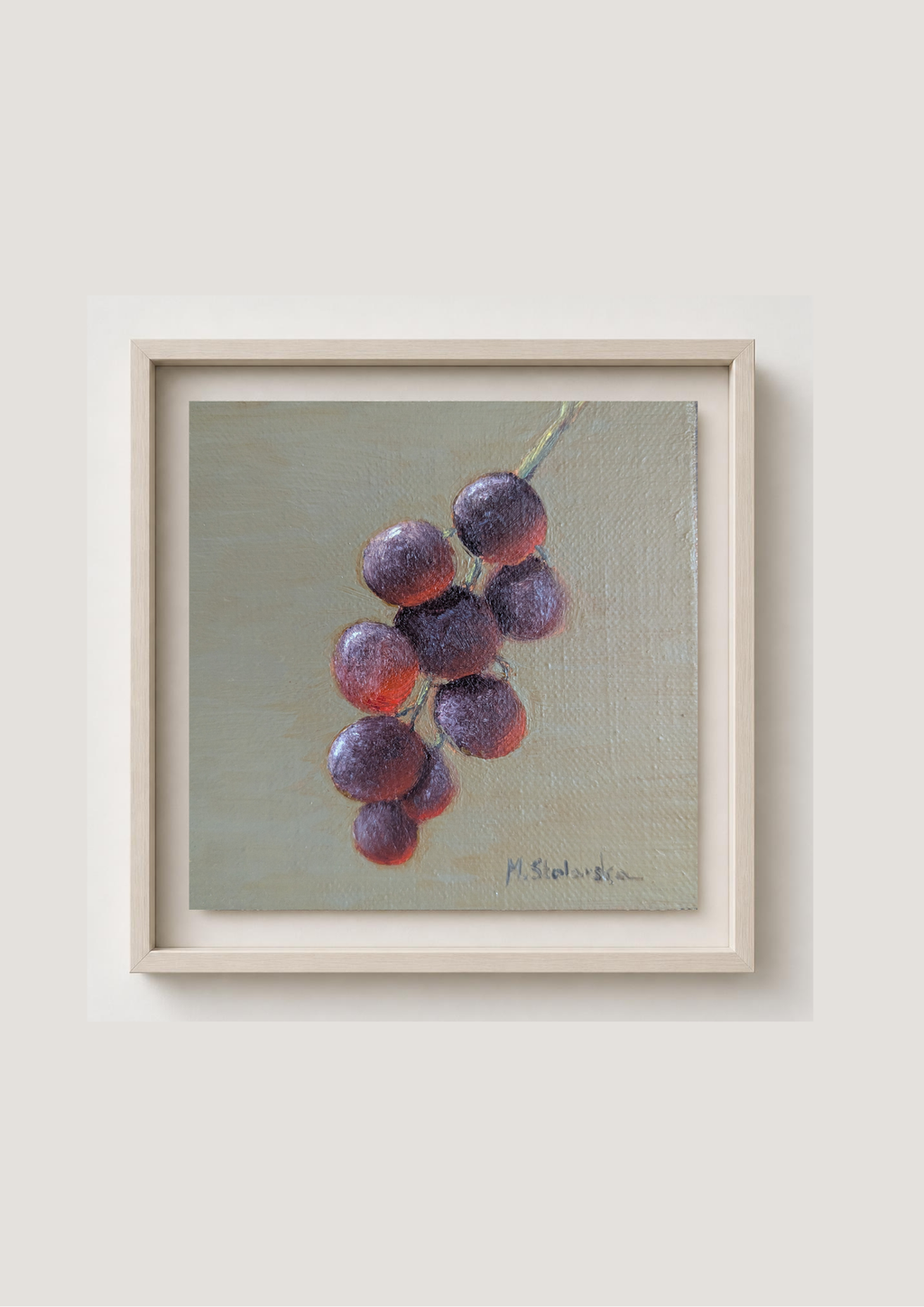 A framed still life painting of a cluster of purple grapes on a muted green-gray background, mounted on a light beige wall.