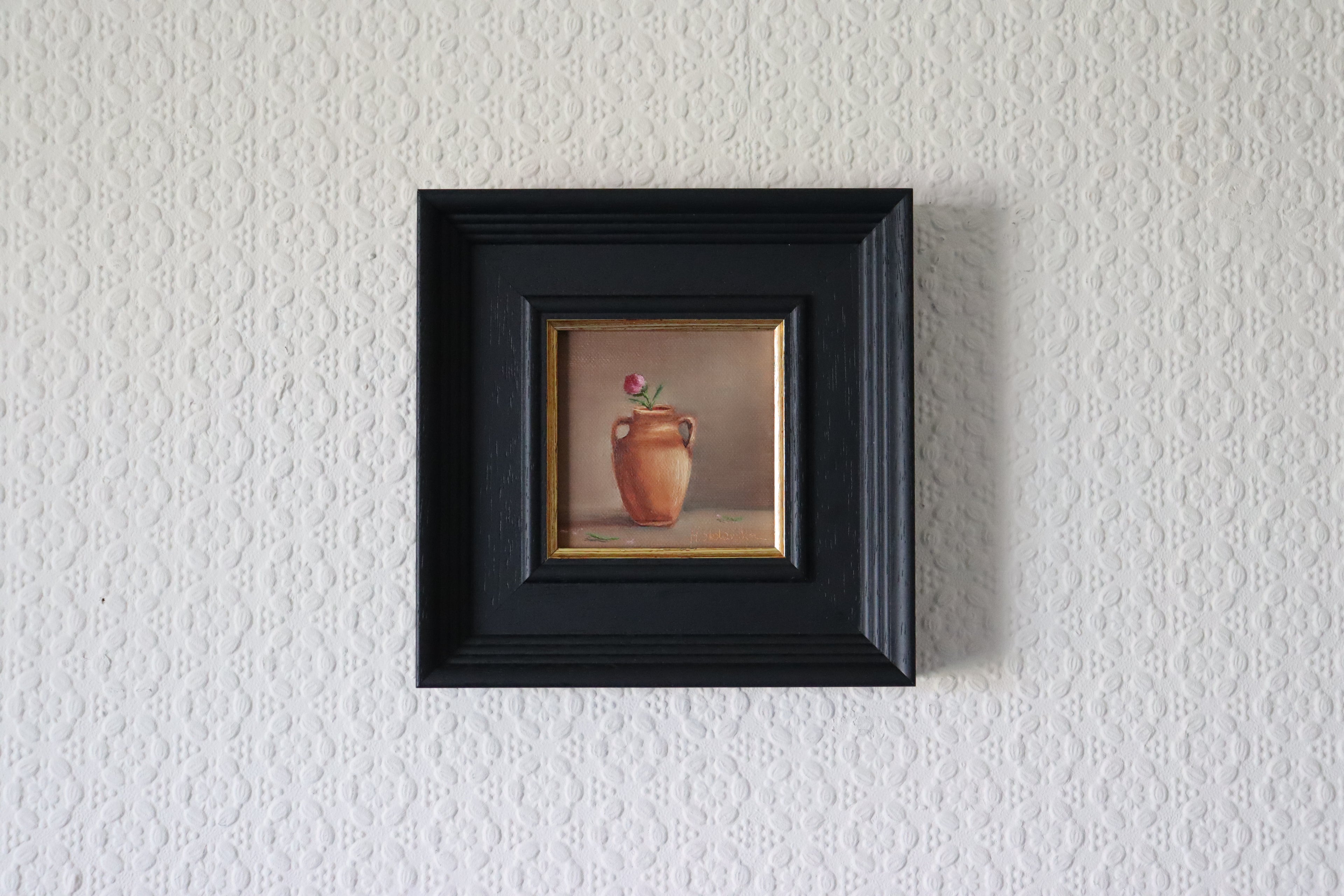 Small painting of a vase with a red flower in a black frame on a textured  Original oil painting of large terracotta glow, warm terracotta vessel in Mediterranean golden lightwhite wall.