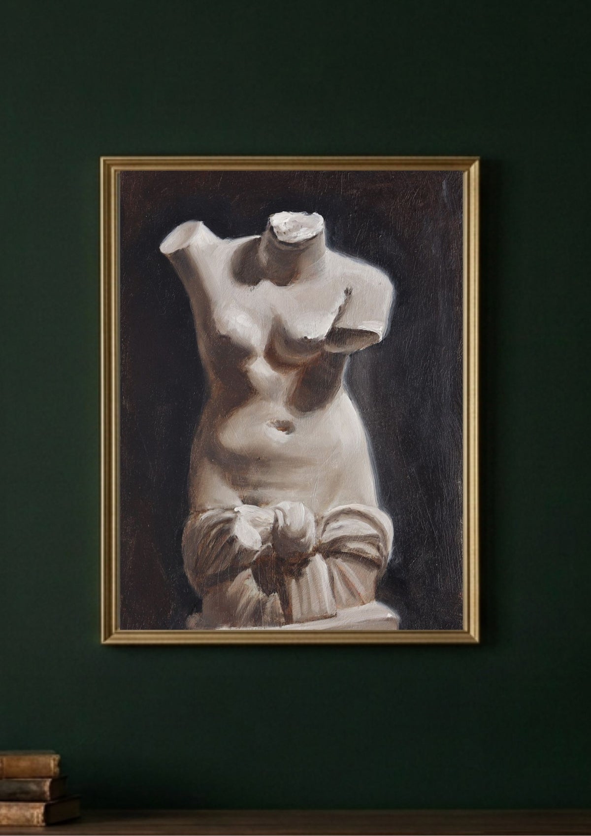 A framed art depicting a classical white marble female torso sculpture against a dark background, hung on a dark green wall above a wooden surface with books.