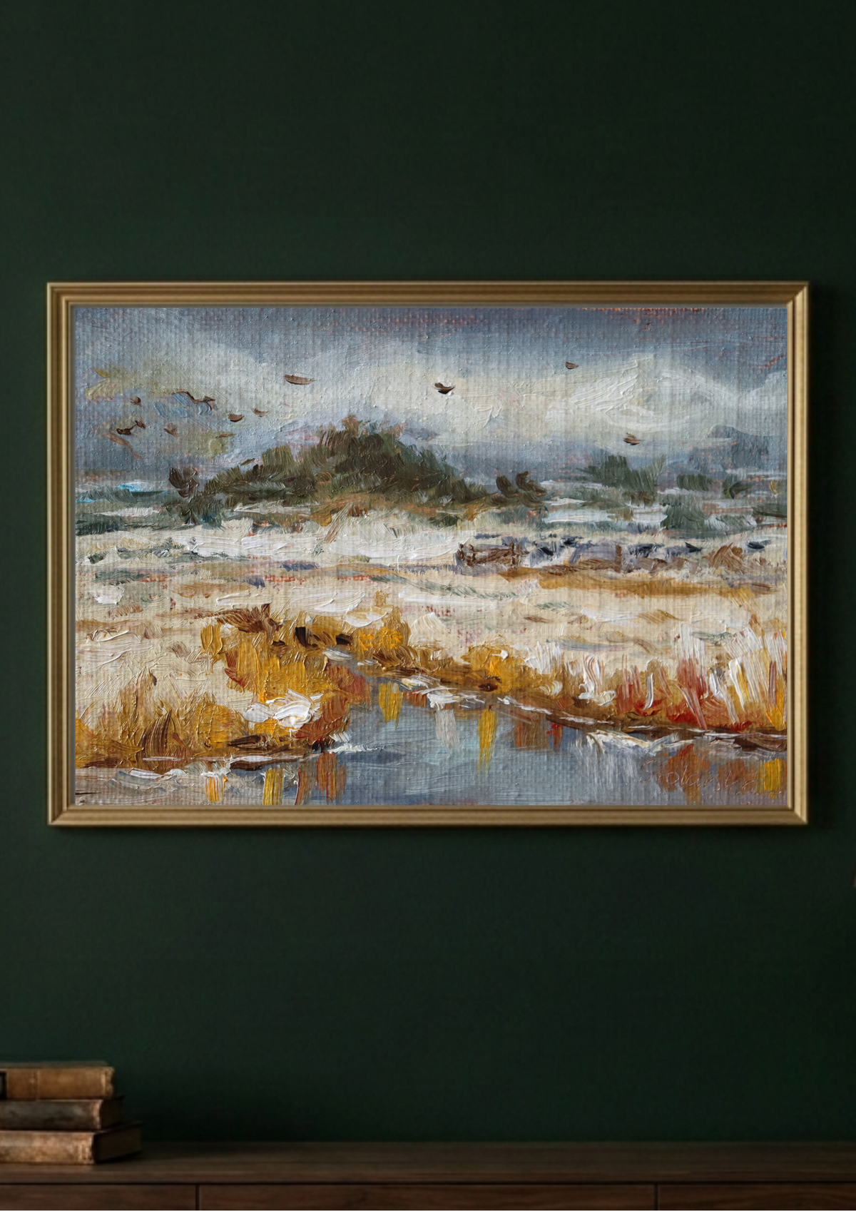 A framed landscape painting hanging on a dark green wall above a wooden console table. The painting depicts a marshland scene with golden reeds, a small stream, distant trees, and birds in muted blue-gray and brown tones, enclosed in a thin gold frame.